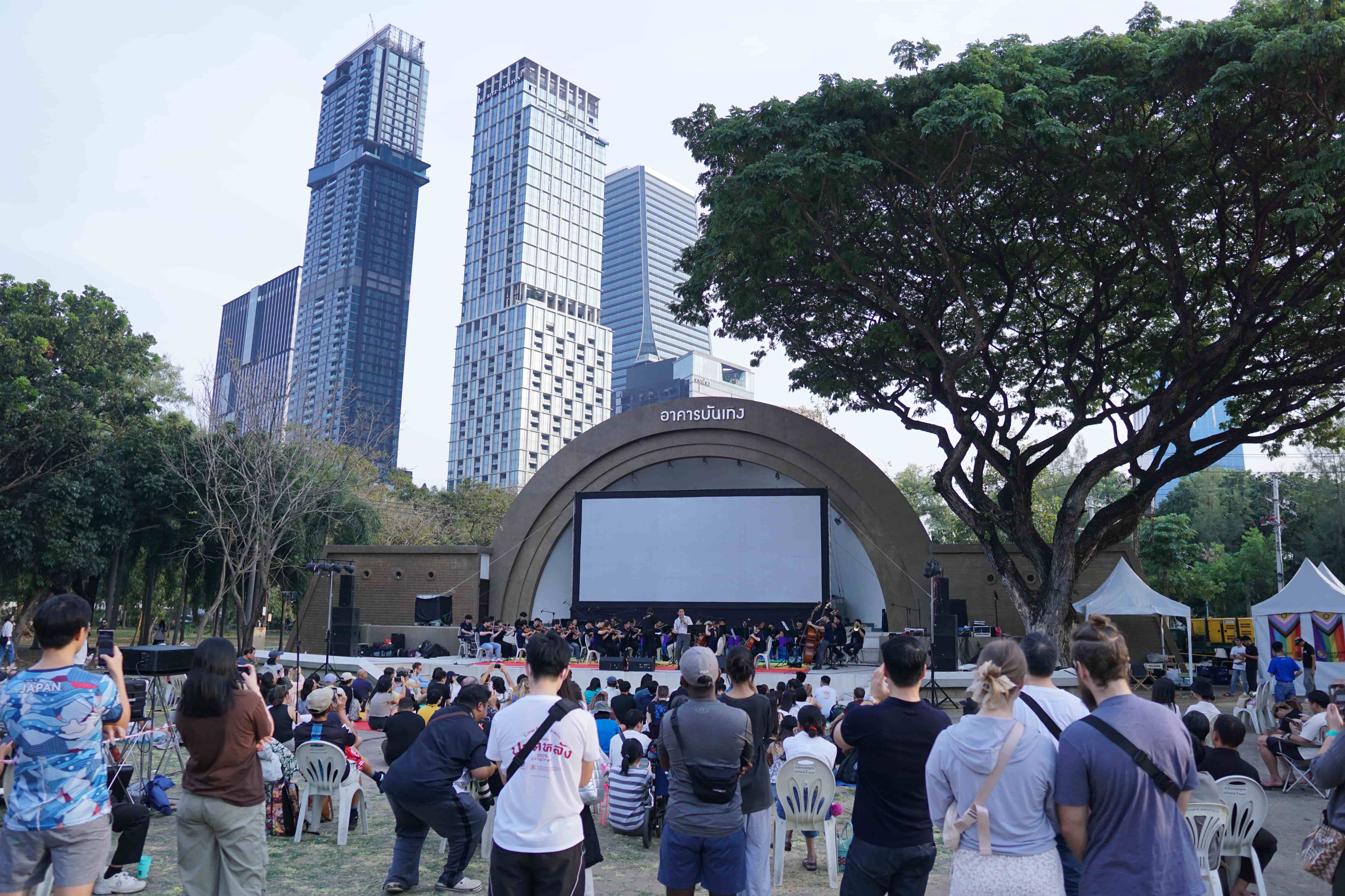 Eine Bühne mit vielen Menschen in einem Park. Zahlreiche weitere Personen von hinten, die stehen oder sitzen und auf die Bühne blicken. Im Hintergrund Hochhäuser.