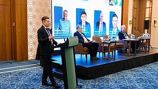 2 Männer sitzen auf Podium, ein weiter steht an Rednerpult