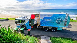 Ein wasserstoffbetriebener Müllwagen mit der Aufschrift „Powered by Hydrogen“ sammelt Müll an einer Küstenstraße mit Blick auf das Meer. Im Vordergrund befinden sich Mülltonnen und grüne Pflanzen, im Hintergrund eine sandige Strandlandschaft.