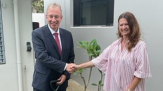 Der deutsche Botschafter Dr. Andreas Prothman und Iris Heinz, AHK Neuseeland, schütteln Hände vor einer kleinen Gedenktafel