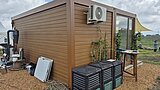 Braunes Containergebäude mit verschiedenen Geräten, drei schwarzen Kompostbehältern davor und einem kleinen Tisch neben einer Glas-Tür auf einem Kiesboden