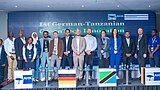 Gruppe von zwölf Personen steht nebeneinander auf einer Bühne vor einer Leinwand mit der Aufschrift '1st German-Tanzanian Green Tech Innovation'