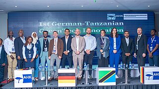 Gruppe von zwölf Personen steht nebeneinander auf einer Bühne vor einer Leinwand mit der Aufschrift '1st German-Tanzanian Green Tech Innovation'