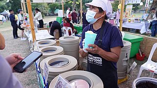 Eine mit Maske und Mütze ausgestattete Frau steht im Freien hinter einem Verkaufsstand und hält einen Becher in der Hand. Im Vordergrund sieht man den Ausschnitt einer Person, die ihr Handy in der Hand hält. 