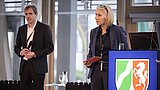 Eine Frau steht an einem Rednerpult mit dem Wappen von Nordrhein-Westfalen. Sie hält ein rotes Tablet in der Hand. Ein Mann steht im Hintergrund mit einem Mikrofon in der Hand. Der Raum hat große Fenster mit Lamellen und eine moderne Einrichtung.