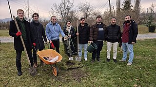 Gruppe von acht Personen steht auf einer Wiese neben einem frisch gepflanzten Baum mit Schubkarre und Gartengeräten