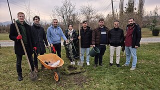 Gruppe von acht Personen steht auf einer Wiese neben einem frisch gepflanzten Baum mit Schubkarre und Gartengeräten