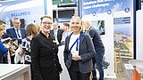 Zwei Frauen stehen in einem Messestandumfeld mit mehreren Personen und Werbetafeln im Hintergrund