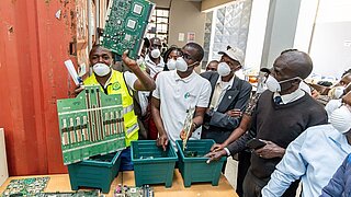 Personen mit Atemmaske stehen vor Boxen, in denen Elektroschrott liegt. Eine Person hält eine Platine in die Höhe. 