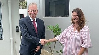 Der deutsche Botschafter Dr. Andreas Prothman und Iris Heinz, AHK Neuseeland, schütteln Hände vor einer kleinen Gedenktafel