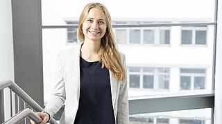 Frau mit langem Haar in weißem Blazer und dunklem Oberteil steht an einem Treppengeländer vor großen Fenstern in einem modernen Gebäude