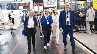 Gruppe von sechs Personen mit Namensschildern geht durch eine Messehalle mit Ständen im Hintergrund