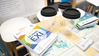 Drei durchsichtige Becher mit schwarzen Deckeln auf einem Tisch neben Broschüren und Flyern mit dem Schriftzug '10 JAHRE EXI'