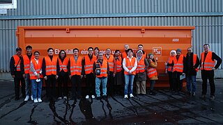 Eine Menschengruppe steht vor einem orangefarbenen Abfallcontainer.