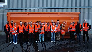 Eine Menschengruppe steht vor einem orangefarbenen Abfallcontainer.