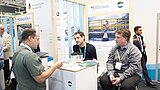 Drei Männer mit Namensschildern sitzen und stehen an einem Messestand mit Informationsmaterial und einem großen Bild im Hintergrund