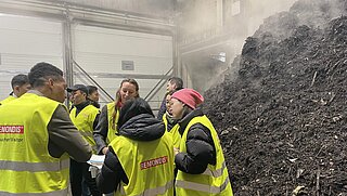 Eine Gruppe von Personen in gelben Westen steht im Kreis über Dokumente gebeugt. Rechts im Bild ist ein großer dampfender Haufen aus Bioabfällen.