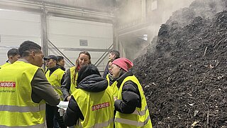 Eine Gruppe von Personen in gelben Westen steht im Kreis über Dokumente gebeugt. Rechts im Bild ist ein großer dampfender Haufen aus Bioabfällen.