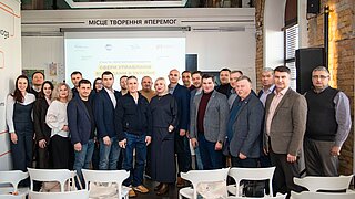 Eine Gruppe von vielen Personen steht in einem Konferenzraum.