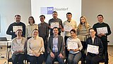 Ein Gruppenfoto von 12 Personen, wovon die Hälfte auf Stühlen sitzt und die andere Hälfte dahinter steht. Mehrere Personen halten ein Zertifikat in der Hand. Im Hintergrund ist eine Folie mit der Aufschrift "IHK Düsseldorf" zu sehen. 