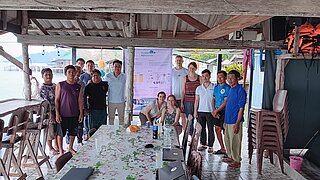 Eine Gruppe von etwa 15 Personen steht in einem offenen, überdachten Bereich in einem Küstendorf. Im Hintergrund hängt ein Poster mit der Aufschrift "Green H2Islands" und technischen Diagrammen.