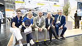 Fünf Personen sitzen nebeneinander vor einem Ausstellungsstück in einer Messehalle mit einem Stand im Hintergrund, der den Schriftzug 'GREEN DRYING SOLUTIONS' trägt