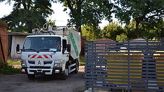 Müllwagen fährt durch kleine Siedlung, in der Abfallcontainer hinter Gittern stehen
