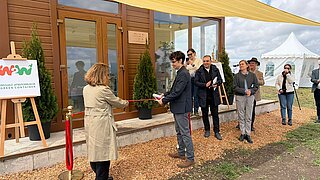 Menschen stehen in einer Reihe vor einem Gebäude mit Glasfront und warten, während eine Frau mit Trenchcoat mit einem Mann mit Sonnenbrille und dunkler Jacke ein rotes Band hält