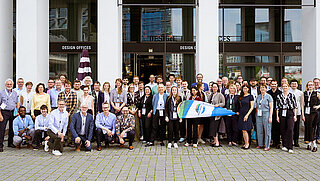 Eine größere Gruppe von Personen steht oder hockt vor einem Gebäude mit weißen Säulen. Vorne halten Personen eine Beach Flag in den Händen. 