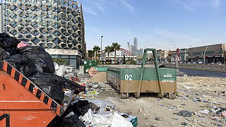 eine Fläche mit mehreren Containern voll Müllsäcken, auf dem Boden liegt unsortierter Müll, im Hintergrund die Stadt Riad