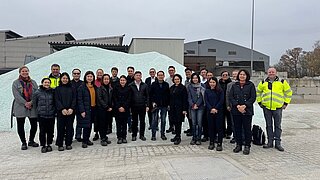 Eine größere Gruppe von Menschen steht auf einem Fabrikgelände vor einem großen Berg aus kleinsten Glasteilen, der hinter ihren Köpfen hervorragt. 