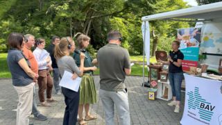 Personen stehen vor Infostand, an dem eine Frau ins Mikro spricht