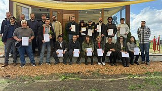 Gruppe von Menschen vor einem Holzgebäude, viele halten Urkunden und einige Blumen, einige sitzen, andere stehen unter einem gelben Sonnensegel