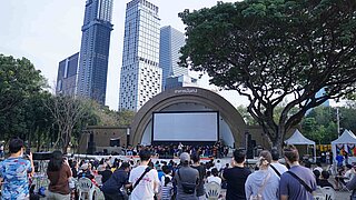 Eine Bühne mit vielen Menschen in einem Park. Zahlreiche weitere Personen von hinten, die stehen oder sitzen und auf die Bühne blicken. Im Hintergrund Hochhäuser.