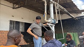 Mehrere Personen stehen bei einem Gebäude, eine Person in grauem Poloshirt und Jeans zeigt auf eine Rohrleitung unter einem Dach