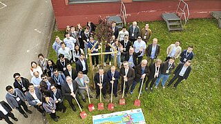 Ein Gruppenfoto von oben auf eine Gruppe von Personen, die auf einer Wiese stehen und Schaufeln in der Halt halten. 