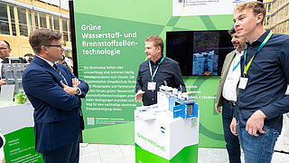 Bundesumweltminister und zwei Männer an einem Stand mit einem Modell eines Containers