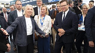 Gruppe von Personen in Businesskleidung auf einer Messe mit einer Frau im Vordergrund, der mit ausgestrecktem Arm spricht